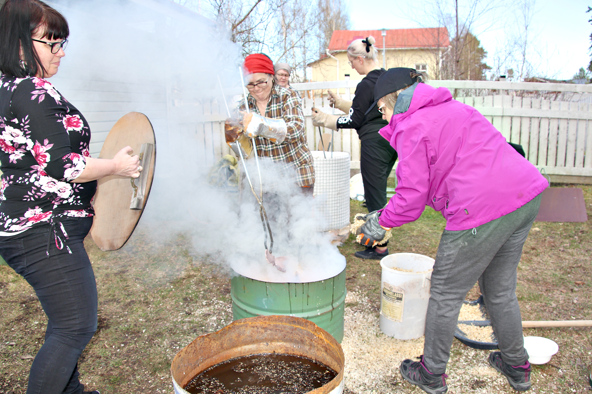 Terhi Miettinen kansi kädessä, Marja Keränen laittaa pihdeillä esineitä savustumaan, Raija Kuosmanen lisää lastuja. Taustalla raku-uunin äärellä Ulla Kortelainen ja Sini Ropponen.