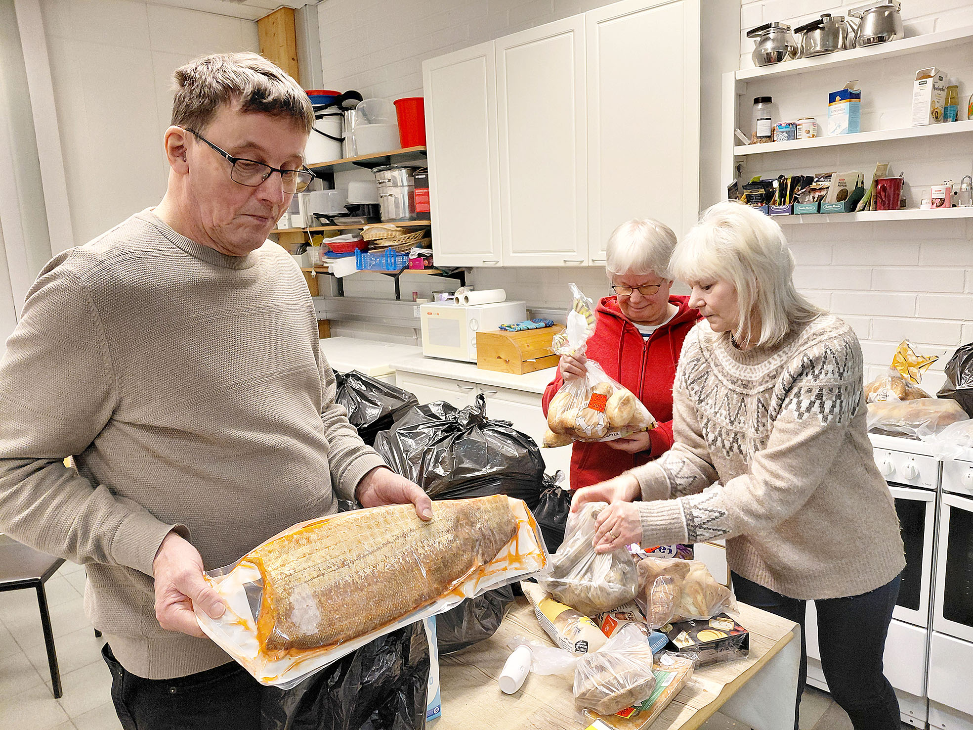 Vapaaehtoiset Urpo Raiskio Kuhmosta, Pirkko Romppanen ja Marja Urpanen Juuasta näyttävät, mitä ruoka-avusta jaettavassa kassissa tällä viikolla on.